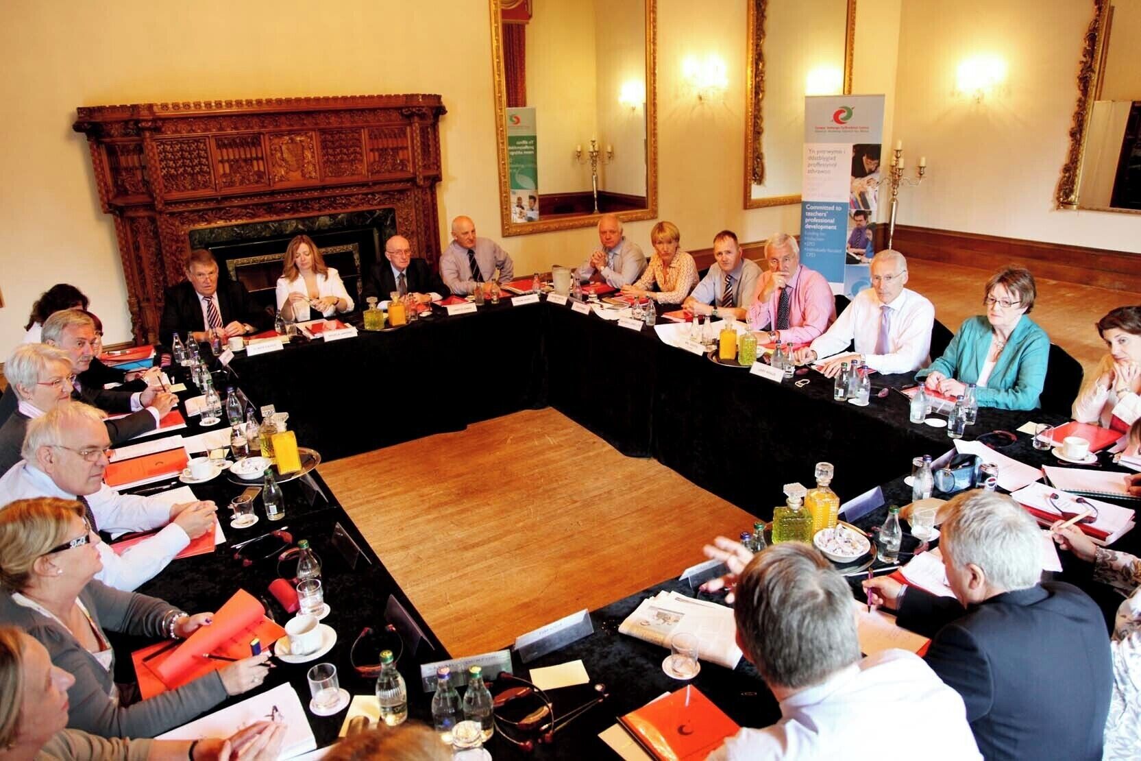 A formal meeting room with a large oval table surrounded by chairs, a fireplace, and a banner on the wall.