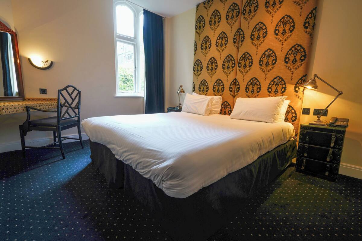 A hotel room with a large bed, a desk with a chair, a window, and a patterned wall.