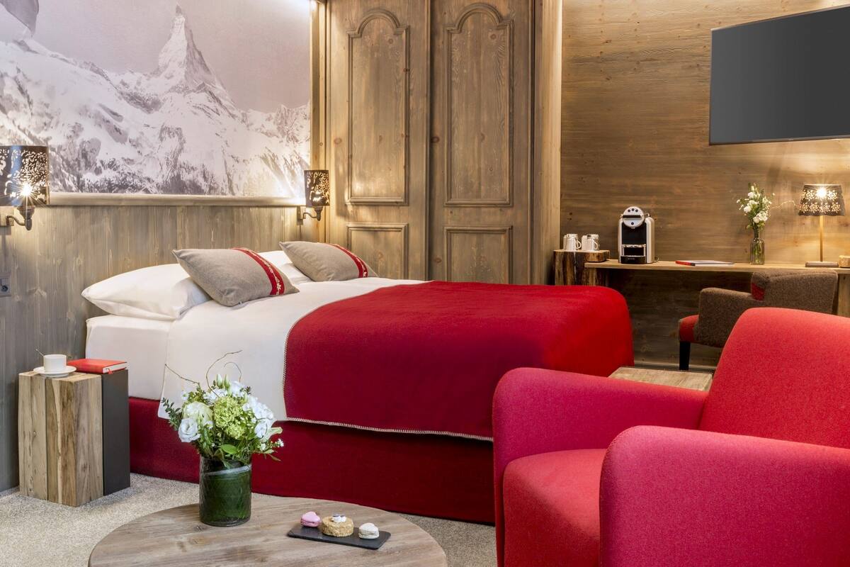 A hotel room with a bed, red upholstered chairs, a wooden desk, and a wall-mounted TV.