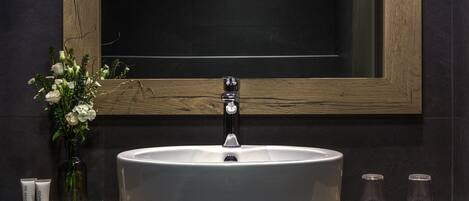 A bathroom with a large white sink, a wooden vanity, a mirror, and a vase with flowers.
