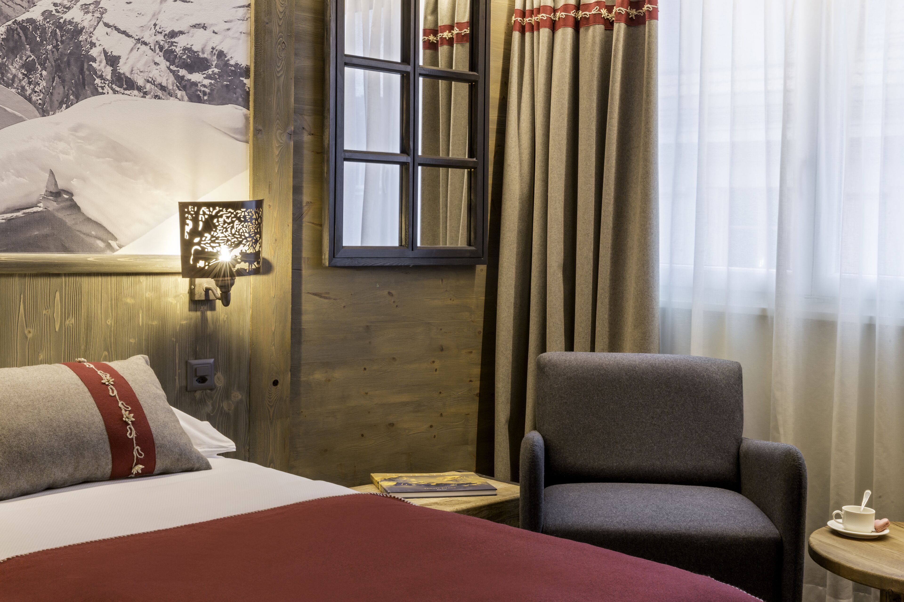 Une chambre d’hôtel avec un lit, une chaise, une petite table avec une tasse, une lampe fixée au mur et un tableau représentant une montagne.