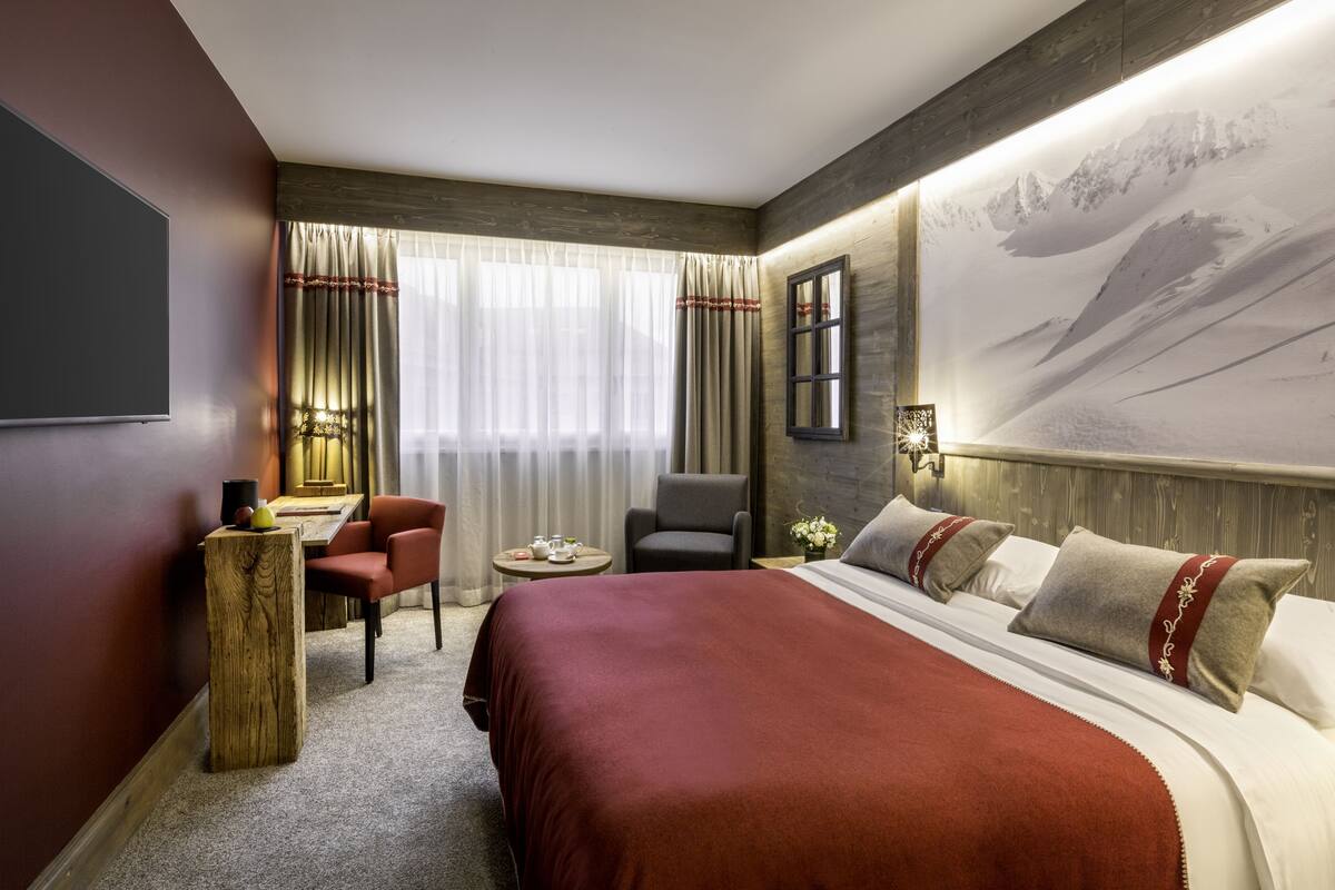 A hotel room with a large bed, a desk with a chair, a small table, and a large window with curtains.