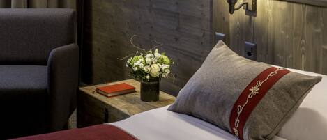 A neatly made bed with a red and grey pillow, a wooden nightstand with a book and a vase of flowers, and a chair.