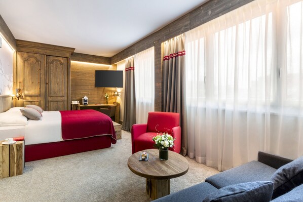 Classic hotel room with a large bed, armchair, and sofa, featuring a flat-screen TV and a small table with a vase of flowers.