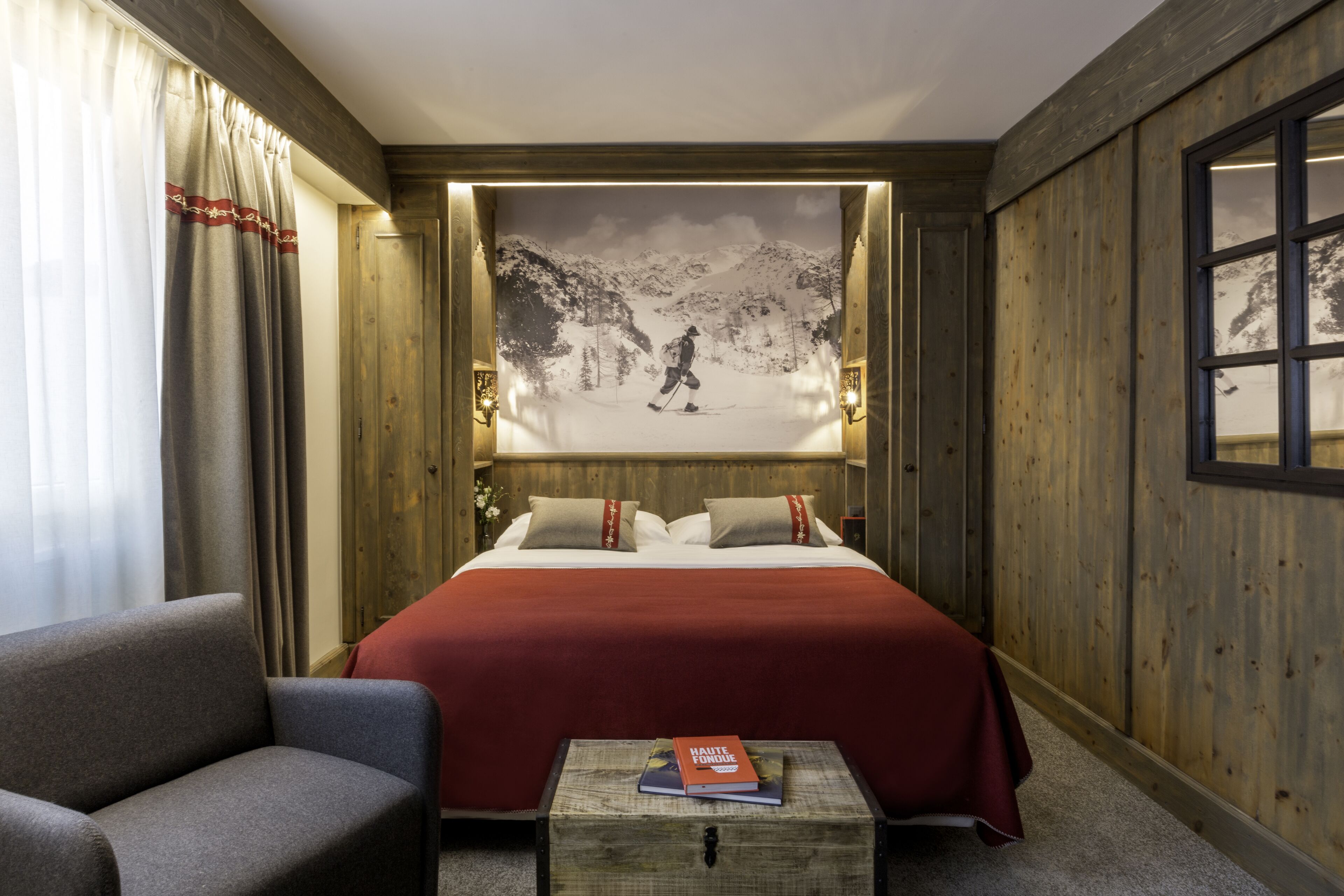 A hotel room with a large bed, a wooden headboard, a small table with a book, and a scenic wall mural.