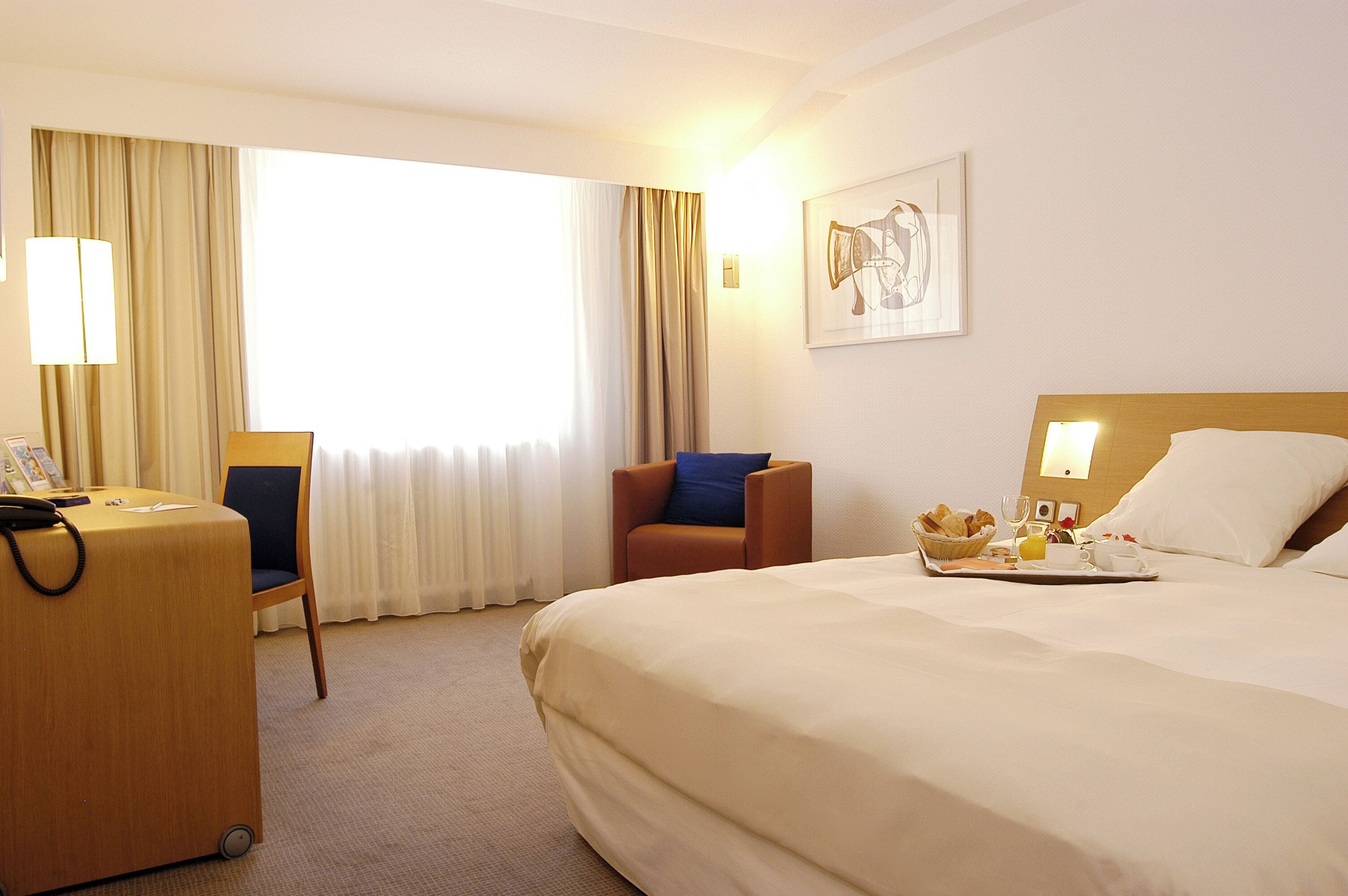 Habitación de hotel con cama, escritorio, silla, ventana con cortinas y una lámpara fijada a la pared.
