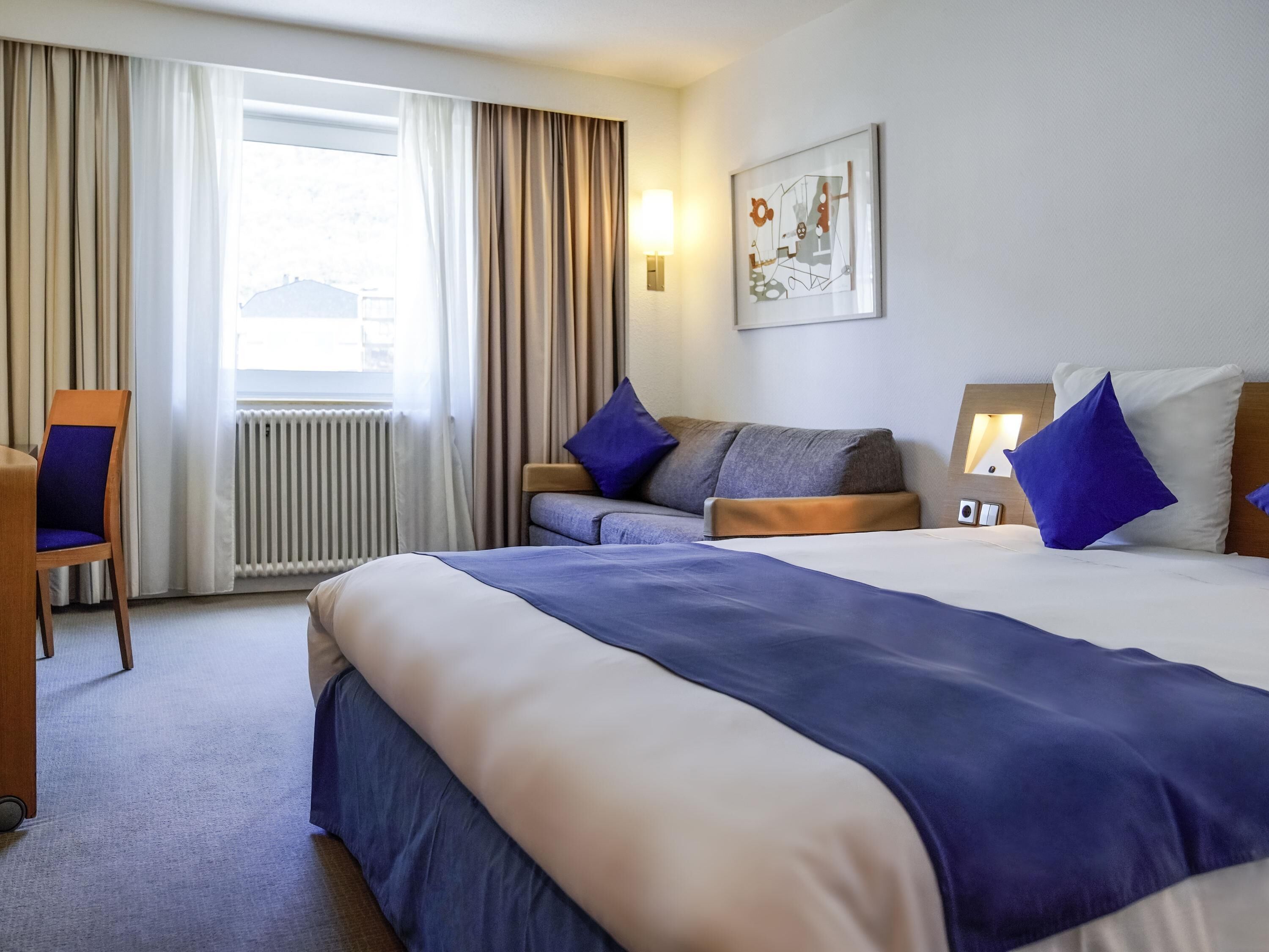 A hotel room with a large bed, a sofa, a chair, and a window with curtains.
