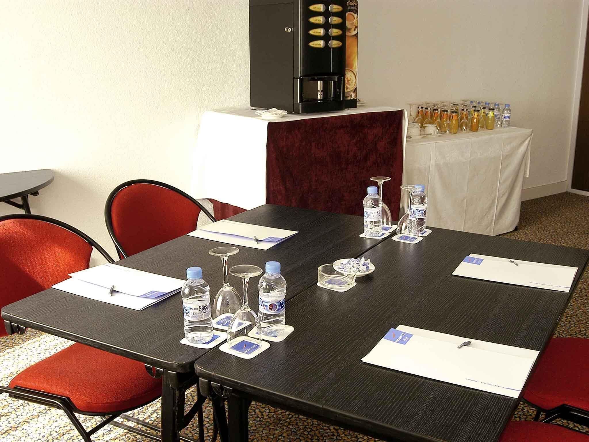 Uma sala de conferências com uma mesa de madeira escura, cadeiras vermelhas, garrafas de água e uma máquina de café.