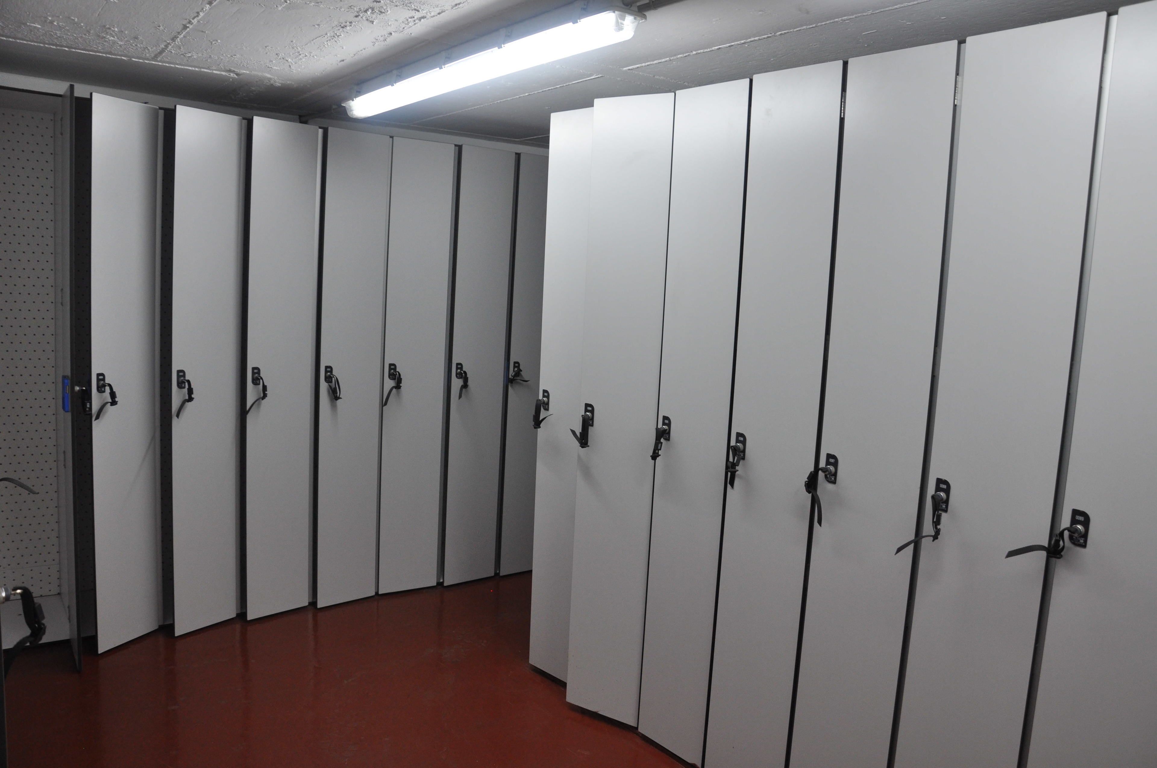 A row of white lockers with metal handles, a red floor, and a fluorescent light fixture on the ceiling.