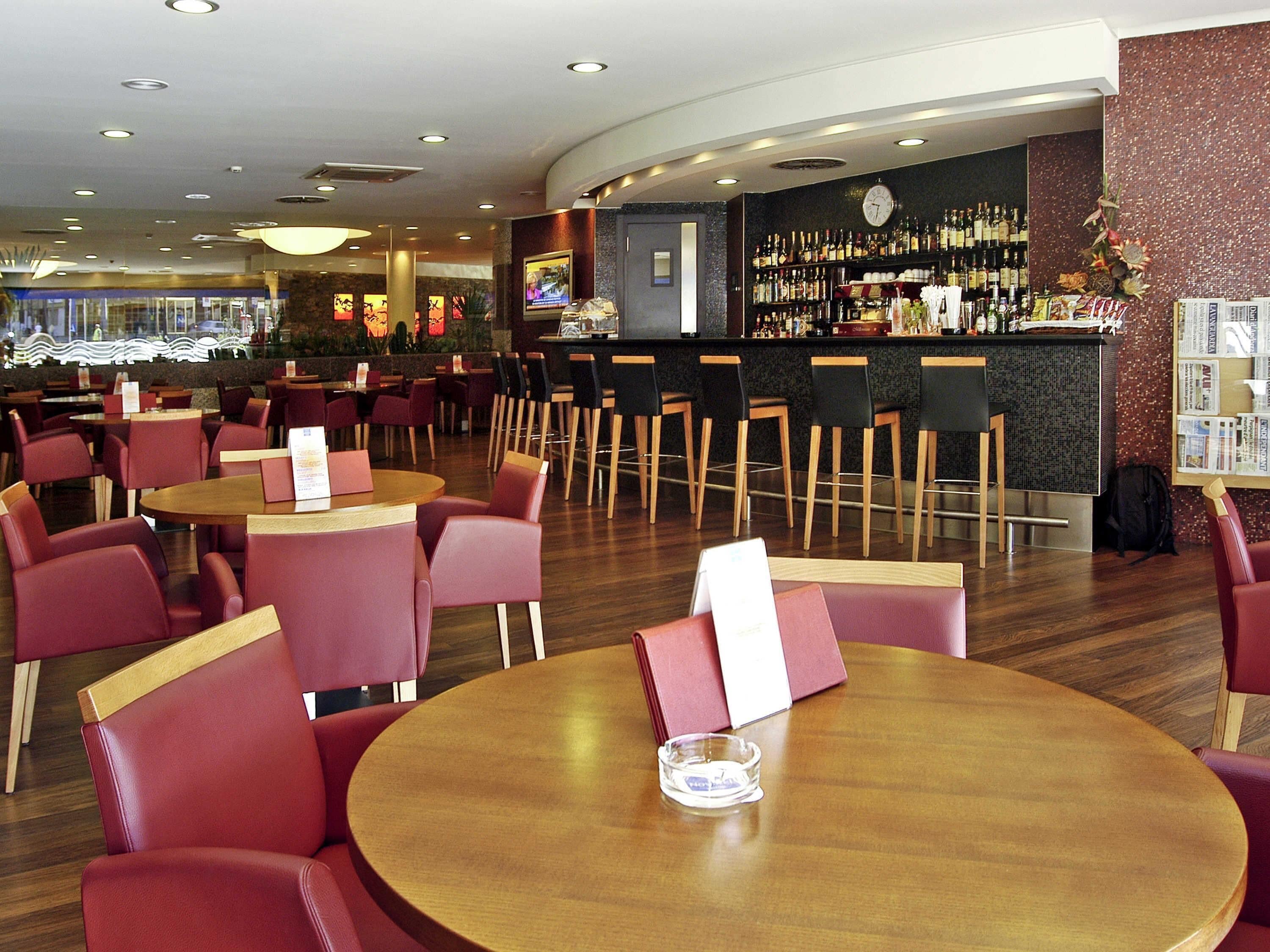 Un restaurante con mesas redondas y sillas rojas, una zona de bar con un reloj y una pantalla de televisión.