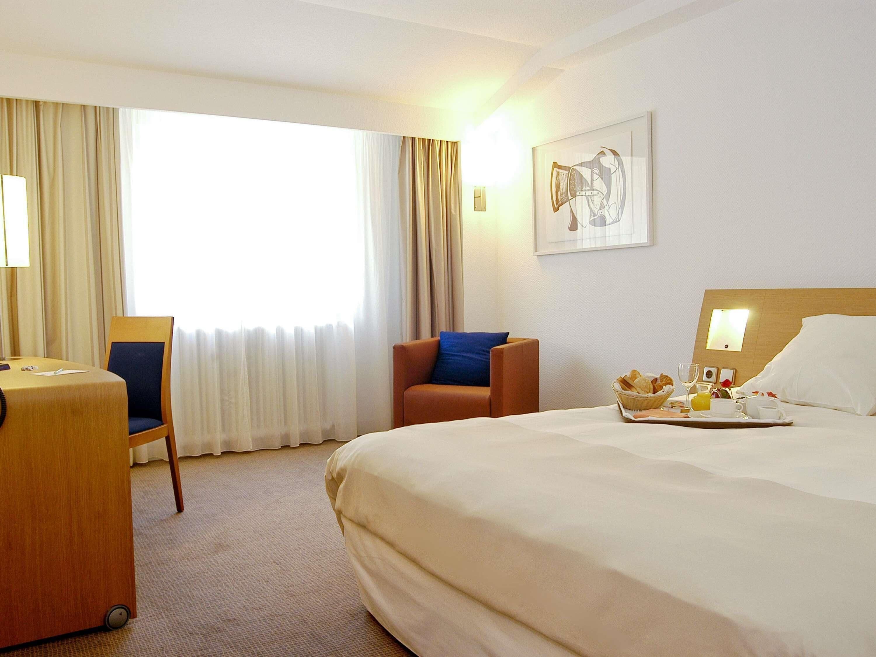 A hotel room with a bed, a desk with a chair, a window with curtains, and a breakfast tray on the bed.