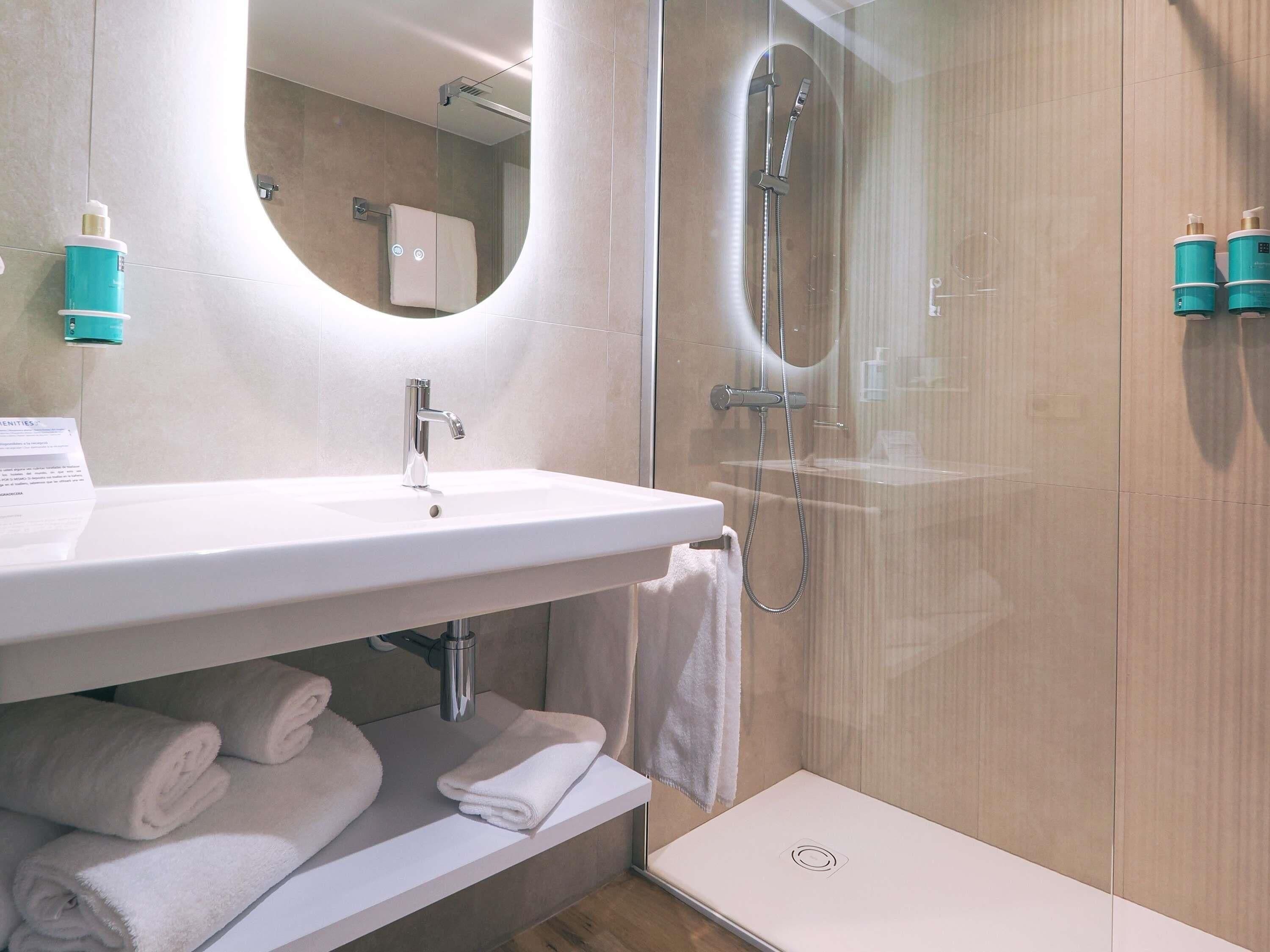 A modern bathroom with a white sink, a glass-enclosed shower, and neatly folded towels on a shelf.