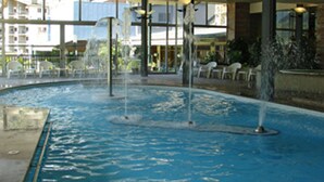 An indoor swimming pool with multiple water jets, surrounded by glass walls and outdoor seating. - Novotel Andorra (Andorra la Vella)