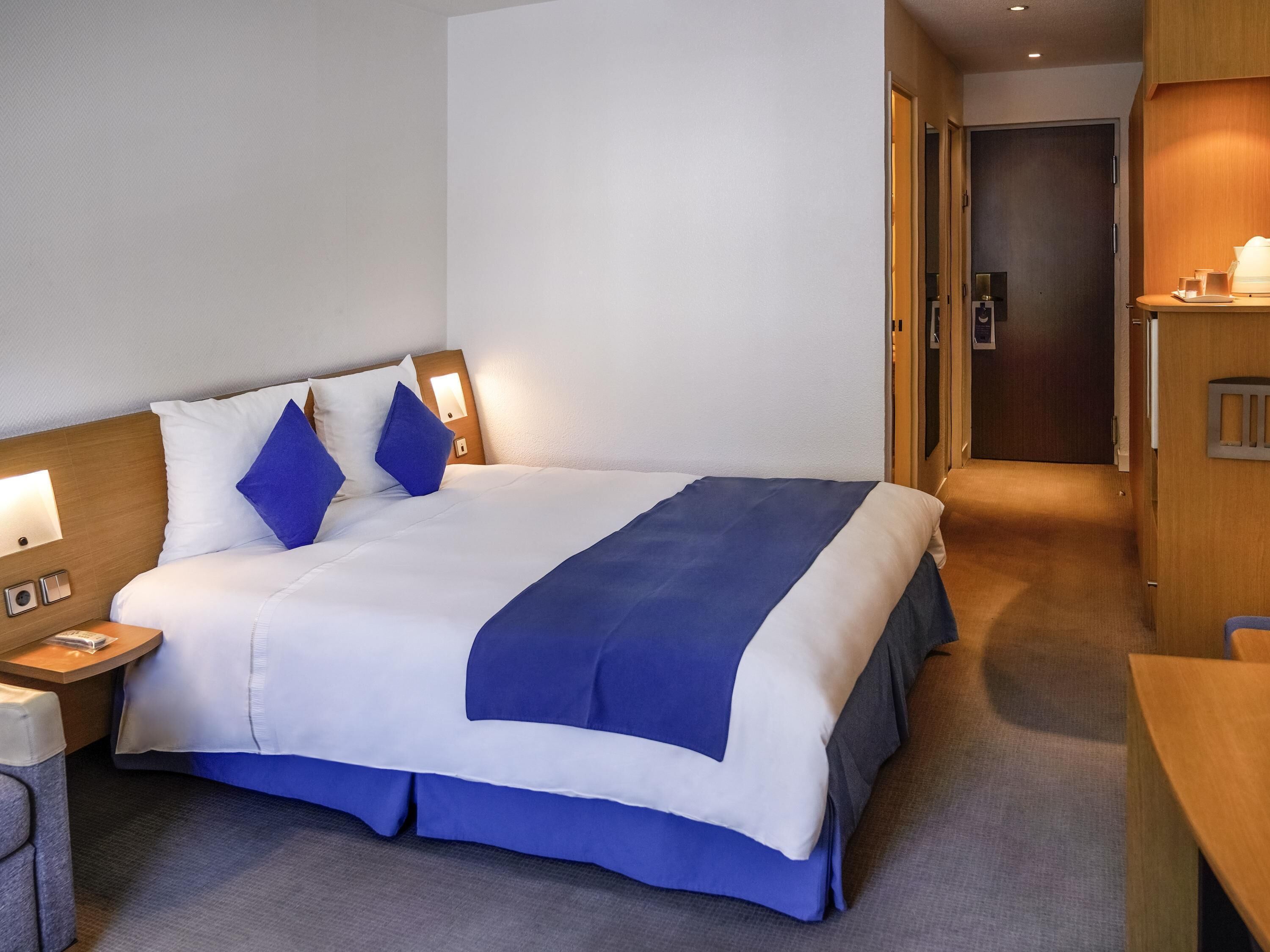 A hotel room with a bed, bedside table, lamp, and a desk with a chair.