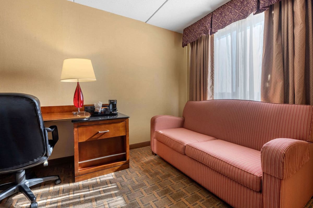 Habitación de hotel con escritorio, lámpara roja, cafetera, sofá rojo y ventana con cortinas.