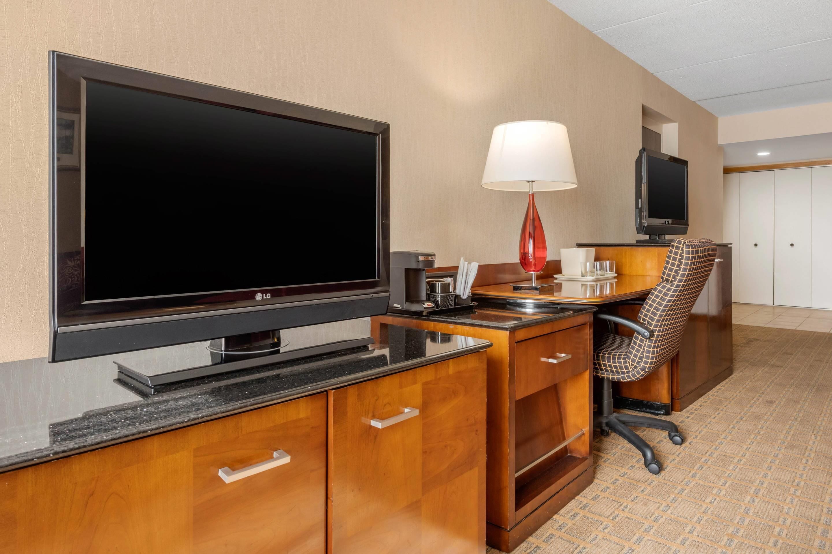 A hotel room with a large flat-screen TV, a desk with a lamp, a patterned office chair, and a wardrobe.