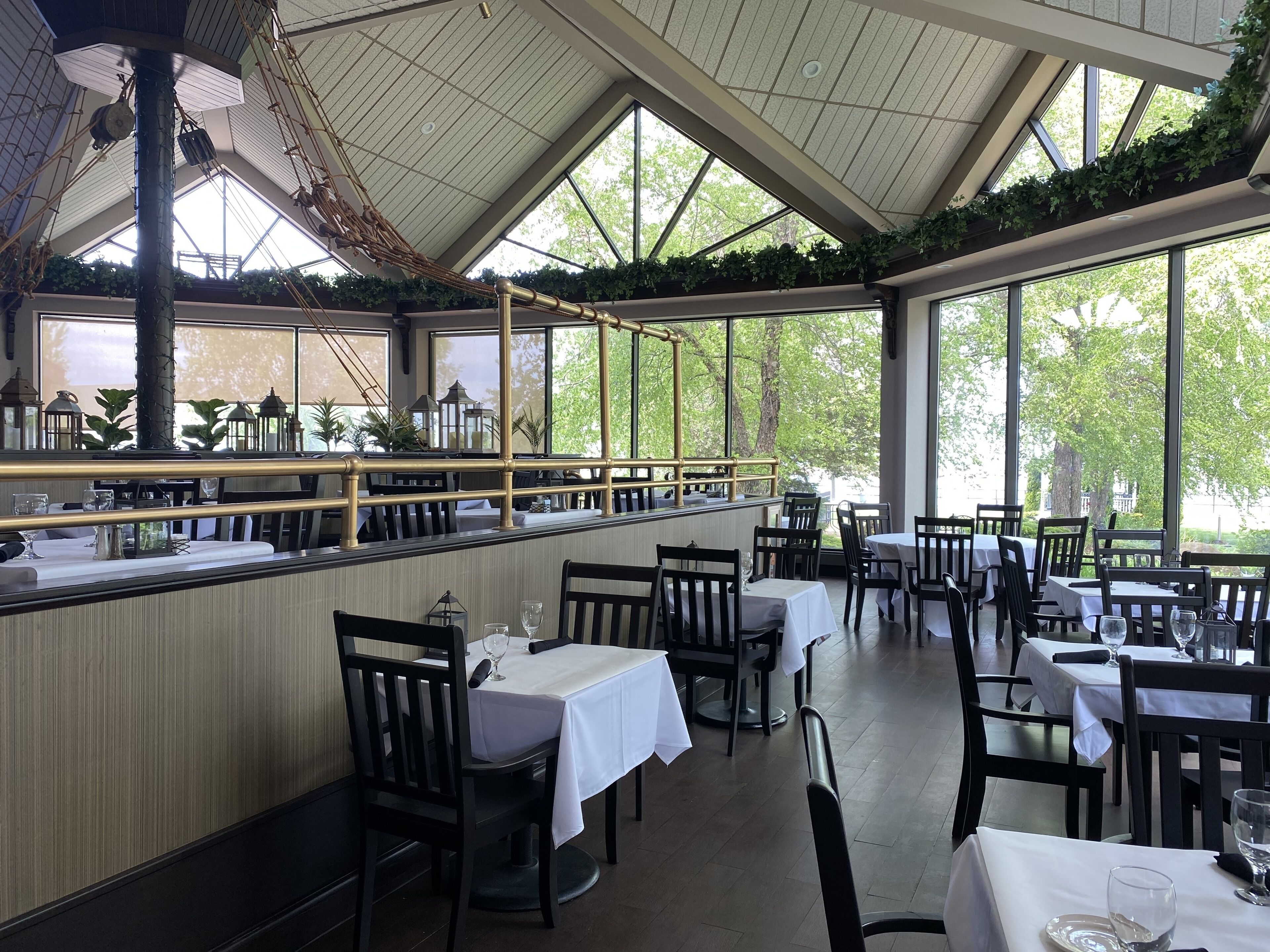 Un restaurante con techo alto, amplios ventanales y vistas al verdor exterior. Las mesas están dispuestas con manteles blancos y sillas negras.