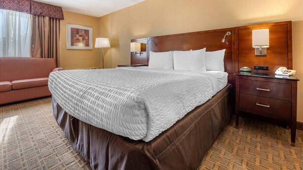 A hotel room with a large bed, a wooden headboard, a bedside table with a lamp, a telephone, and a brown leather sofa.