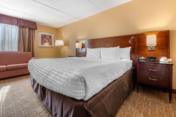 A hotel room with a large bed, a wooden headboard, a bedside table with a lamp, a telephone, and a brown leather sofa. - Clarion Hotel Conference Center on Lake Erie (Dunkirk)