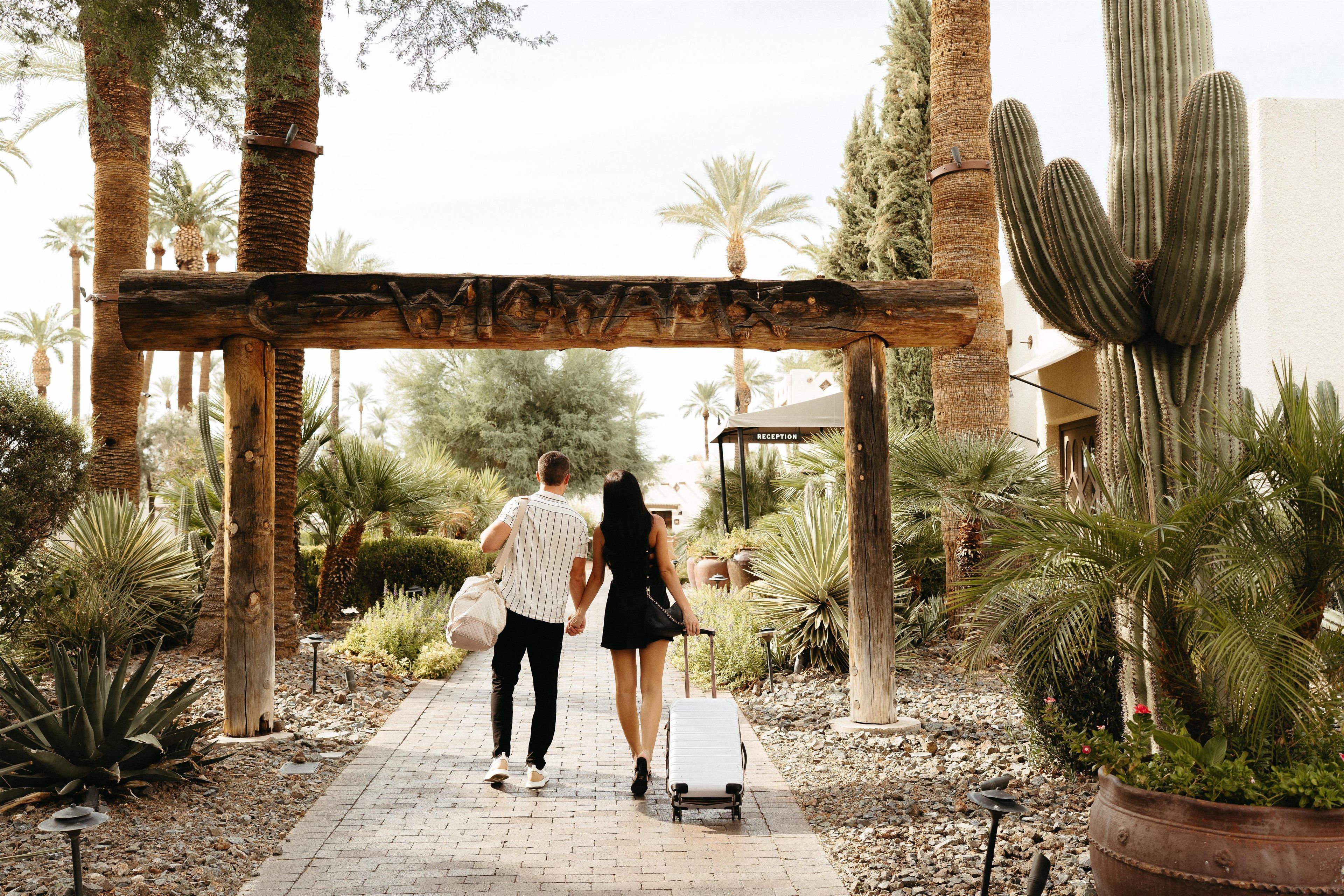 A couple walking under a wooden archway with the word 'RECEPTION' visible, surrounded by desert plants and cacti.