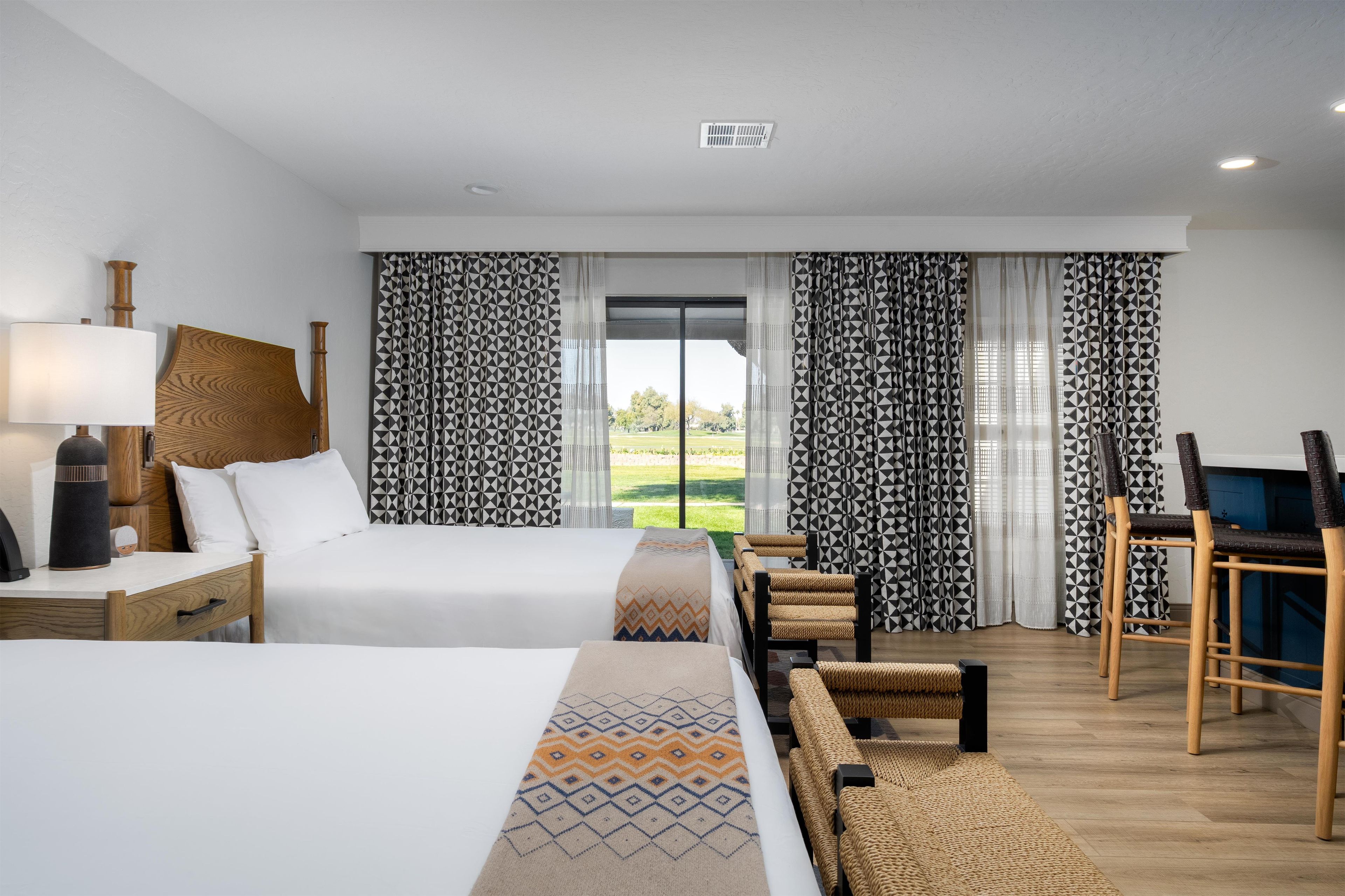 A hotel room with a bed, bedside table, lamp, and a view of the outdoors through the window.