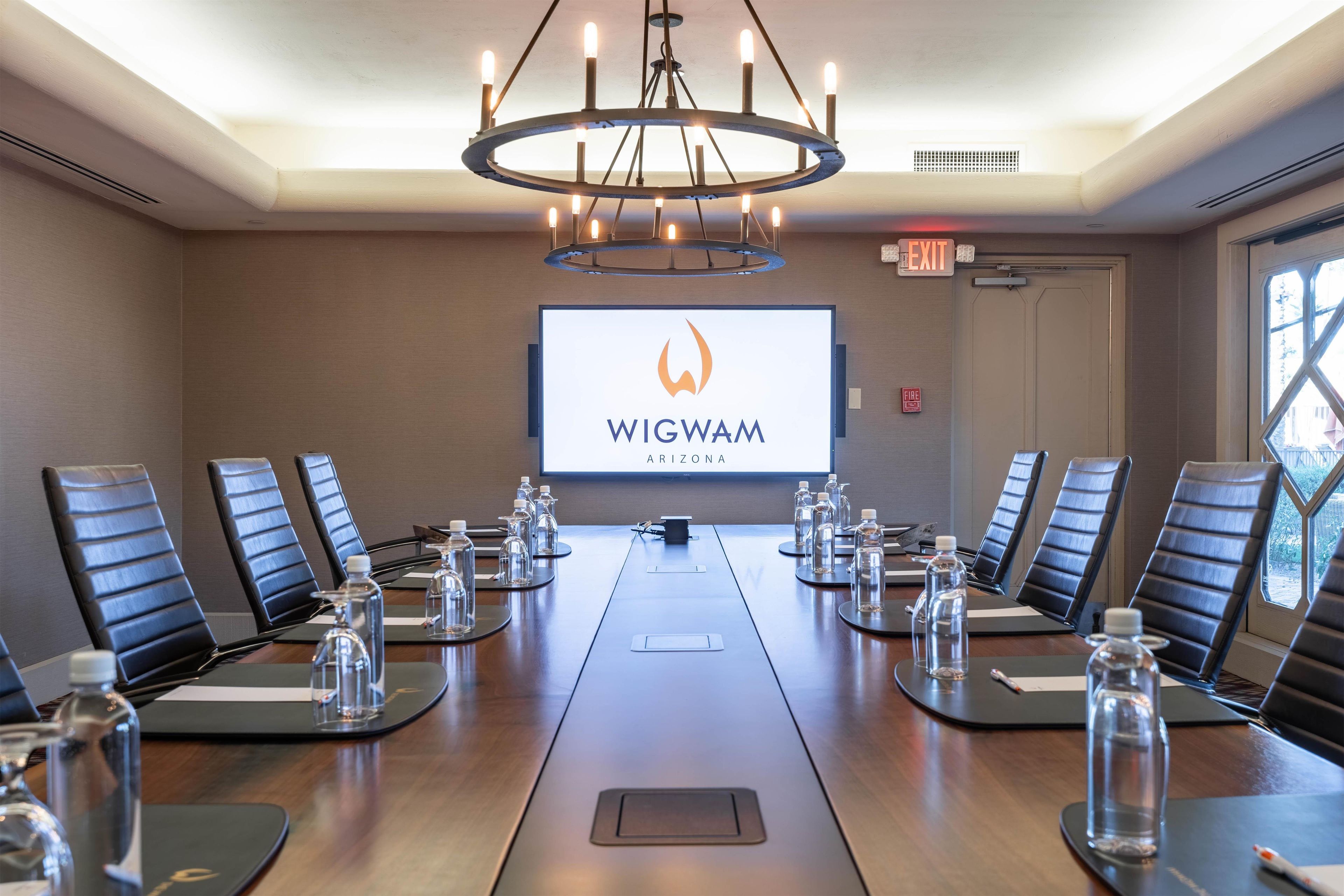 A conference room with a long table, chairs, a projector screen displaying 'WIGWAM ARIZONA', and a chandelier.