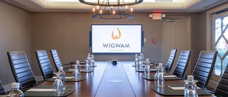 Une salle de conférence avec une longue table, des chaises, un écran de projection affichant le texte « WIGWAM ARIZONA » et un lustre.