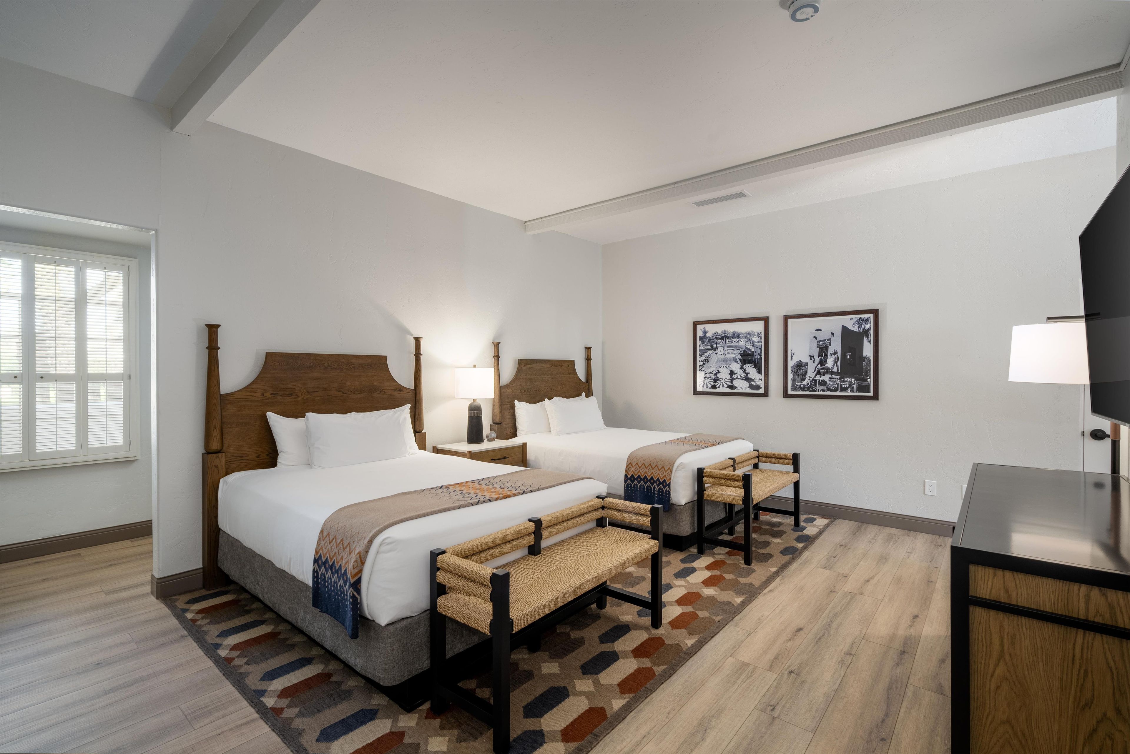 A hotel room with two beds, a desk, a chair, a lamp, and two framed pictures on the wall.