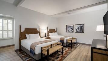 A hotel room with two beds, a desk, a chair, a lamp, and two framed pictures on the wall.
