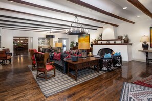 A spacious living room with a fireplace, wooden flooring, and a high ceiling with exposed beams. - The Wigwam (Litchfield Park)