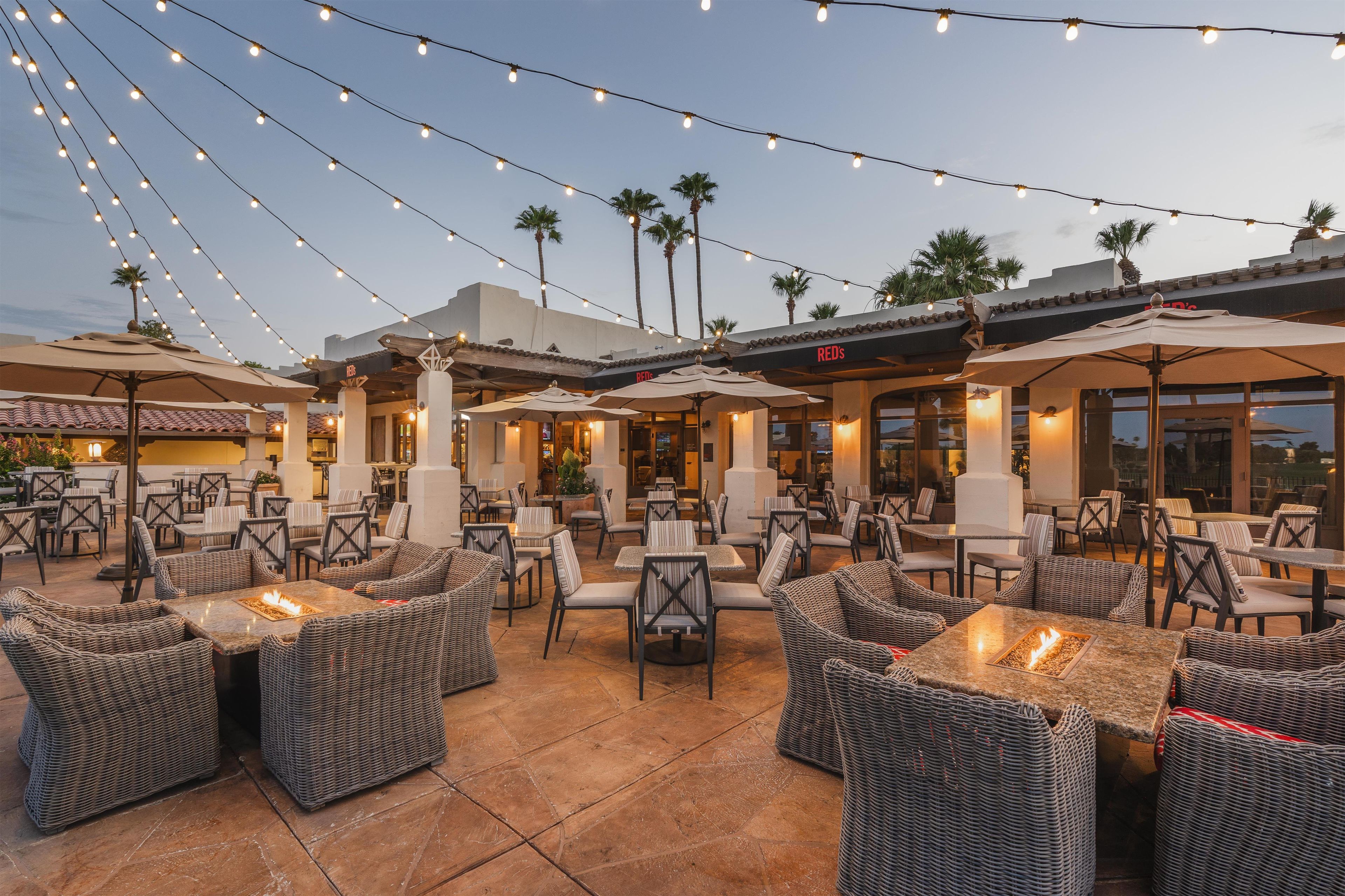 An outdoor patio area with wicker furniture, tables, and chairs, featuring string lights overhead.