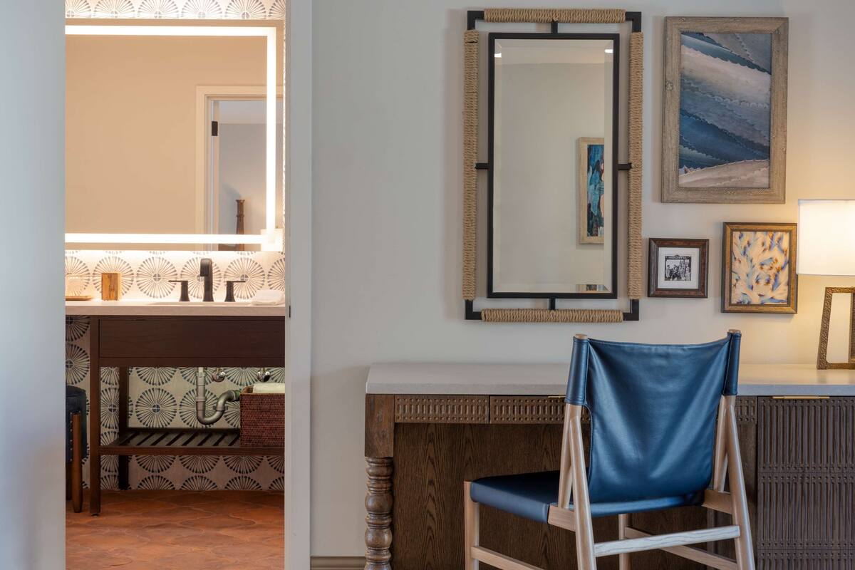 A bathroom with a sink, mirror, and towel rack. There is a chair and a desk with a lamp.