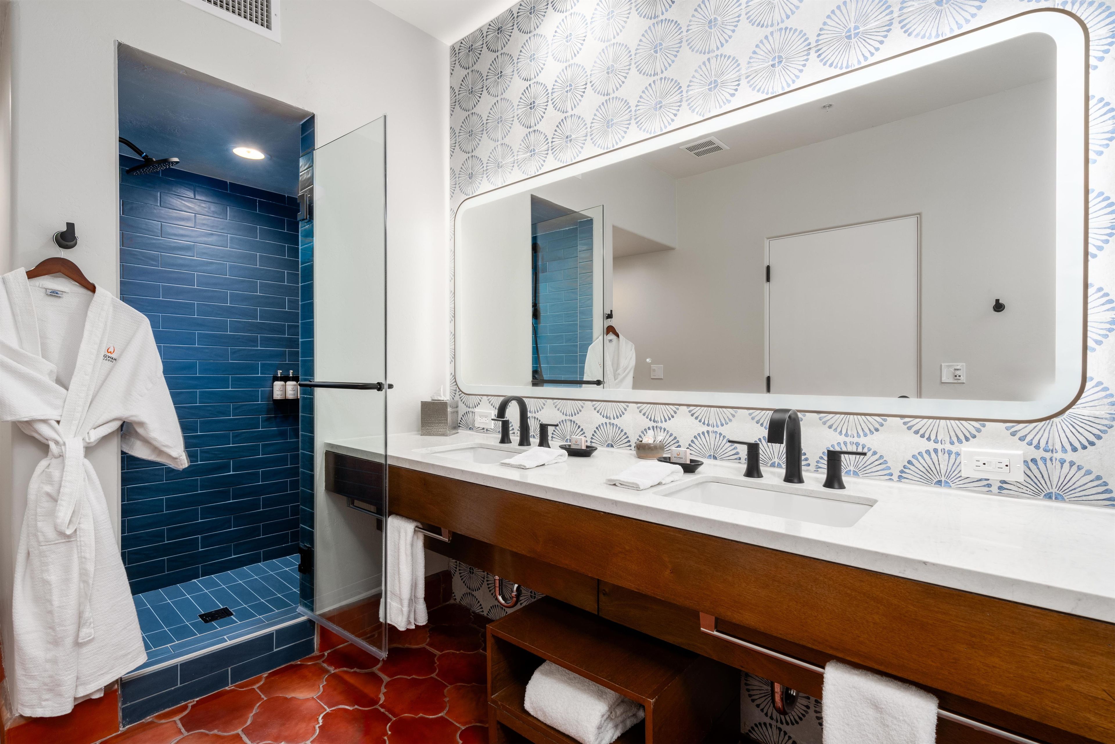 Une salle de bain avec une cabine de douche bleue, une robe blanche suspendue, un grand miroir et un meuble-lavabo en bois doté de deux vasques.