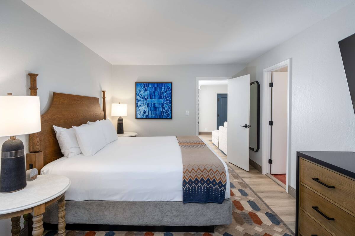 A hotel room with a large bed, bedside table, lamp, and a blue artwork on the wall.