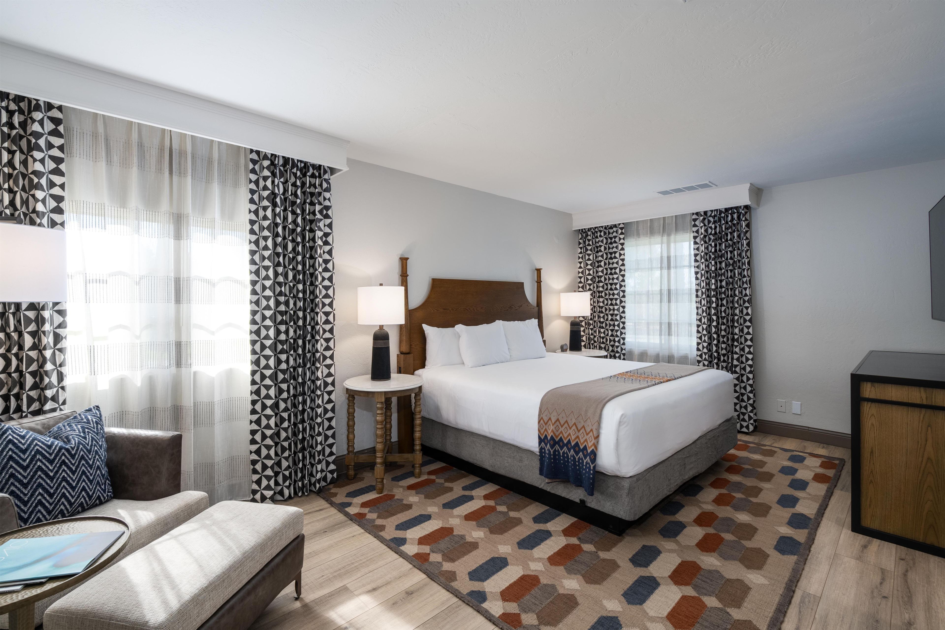 A hotel room with a bed, bedside lamps, a desk, a chair, and patterned curtains.