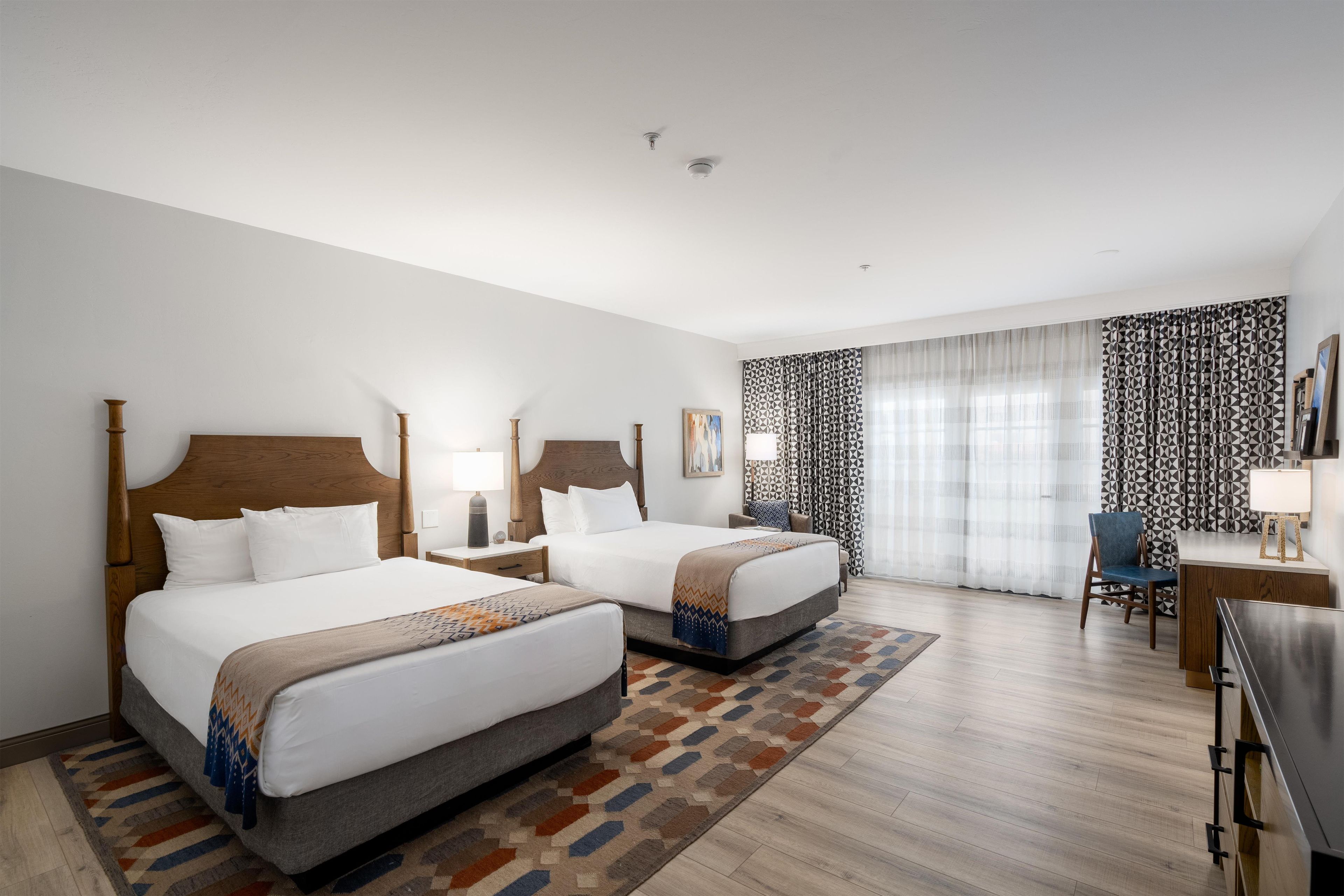 Ein Hotelzimmer mit zwei Betten, einem Schreibtisch, einem Stuhl und einem großen Fenster mit gemusterten Vorhängen.