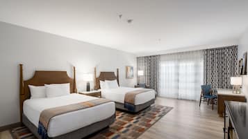 Ein Hotelzimmer mit zwei Betten, einem Schreibtisch, einem Stuhl und einem großen Fenster mit gemusterten Vorhängen.