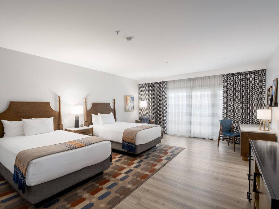 A hotel room with two beds, a desk, a chair, and a large window with patterned curtains.