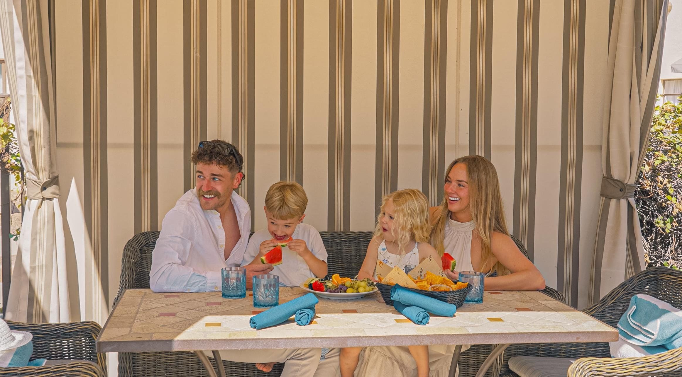 A family dining at an outdoor table with striped curtains in the background.