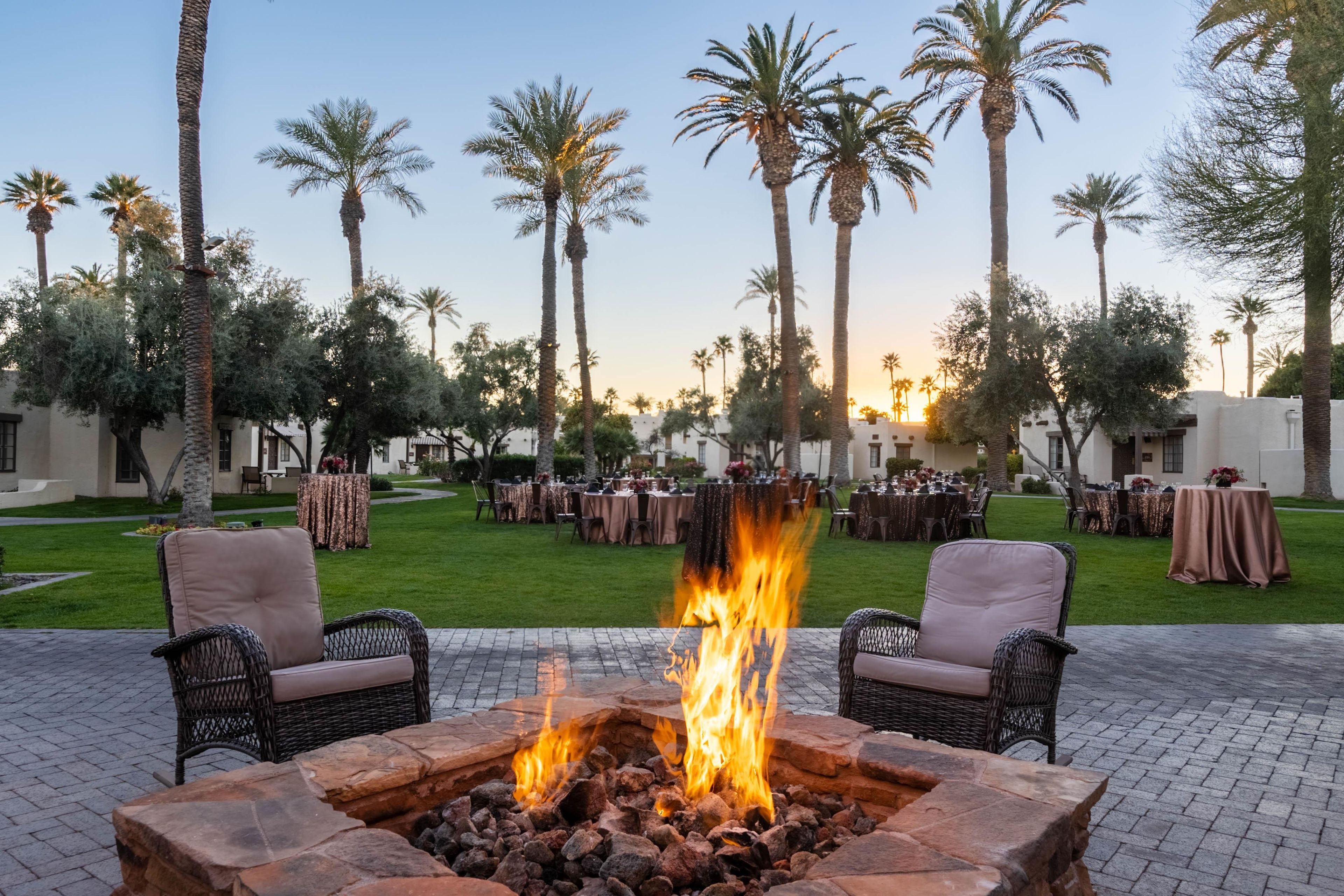 An outdoor seating area with a fire pit, surrounded by palm trees and tables set for dining.