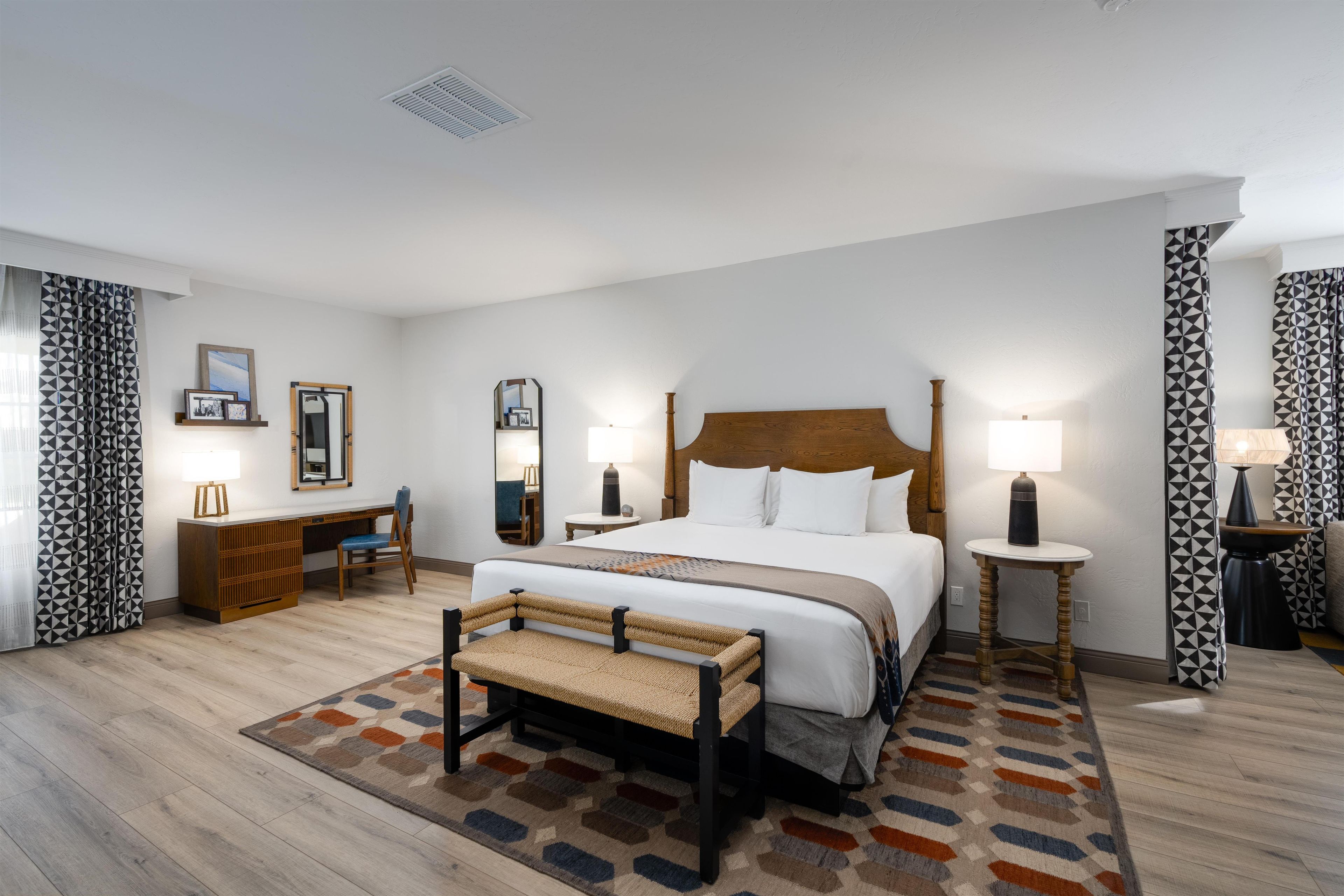 A hotel room with a large bed, bedside tables, a desk, a chair, and patterned curtains.