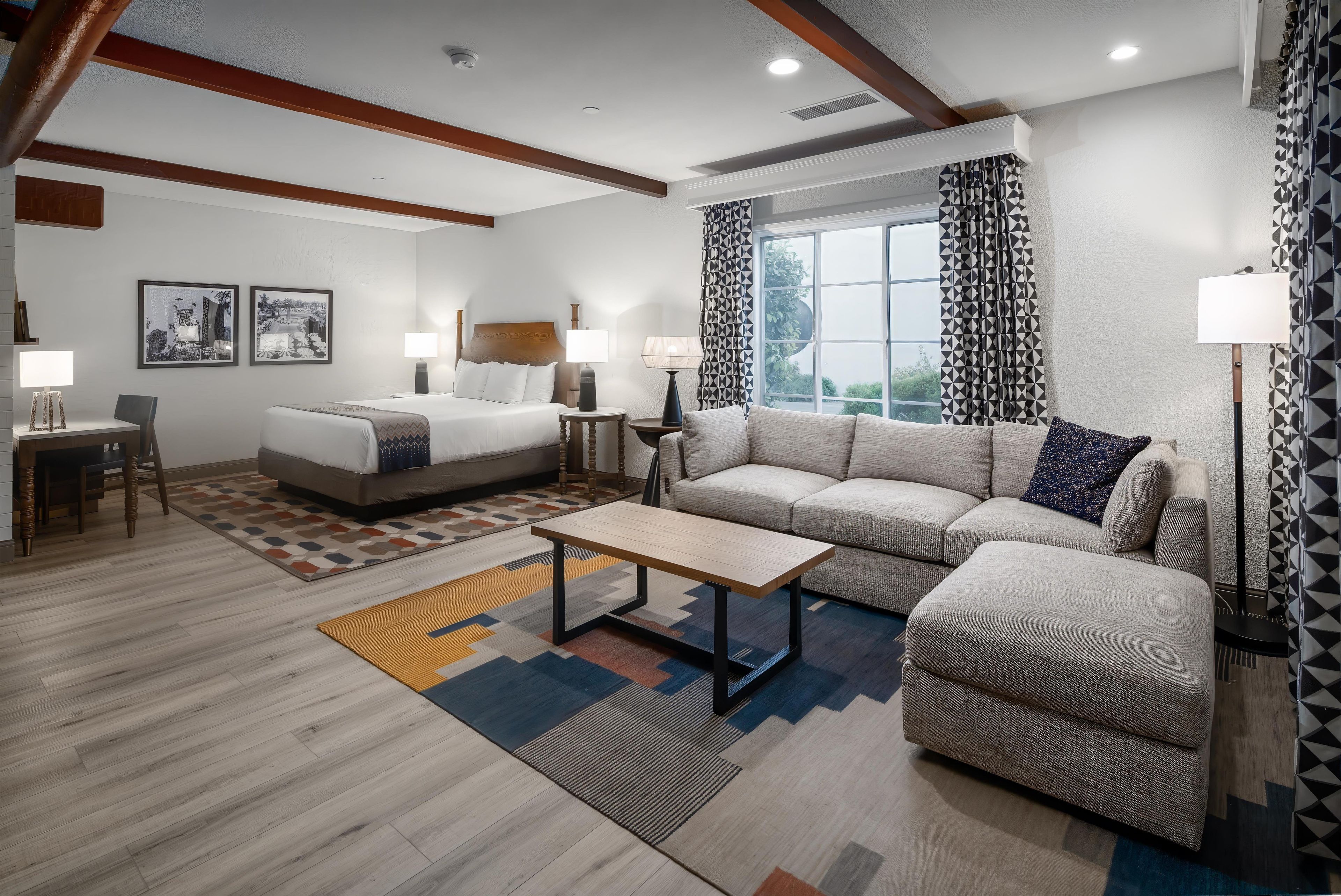 A hotel room with a bed, sofa, and a small table. There are two bedside tables with lamps, a window with curtains, and a patterned rug on the floor.
