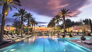 Zona de piscina con sillones de descanso y palmeras, rodeada de edificios con iluminación cálida.