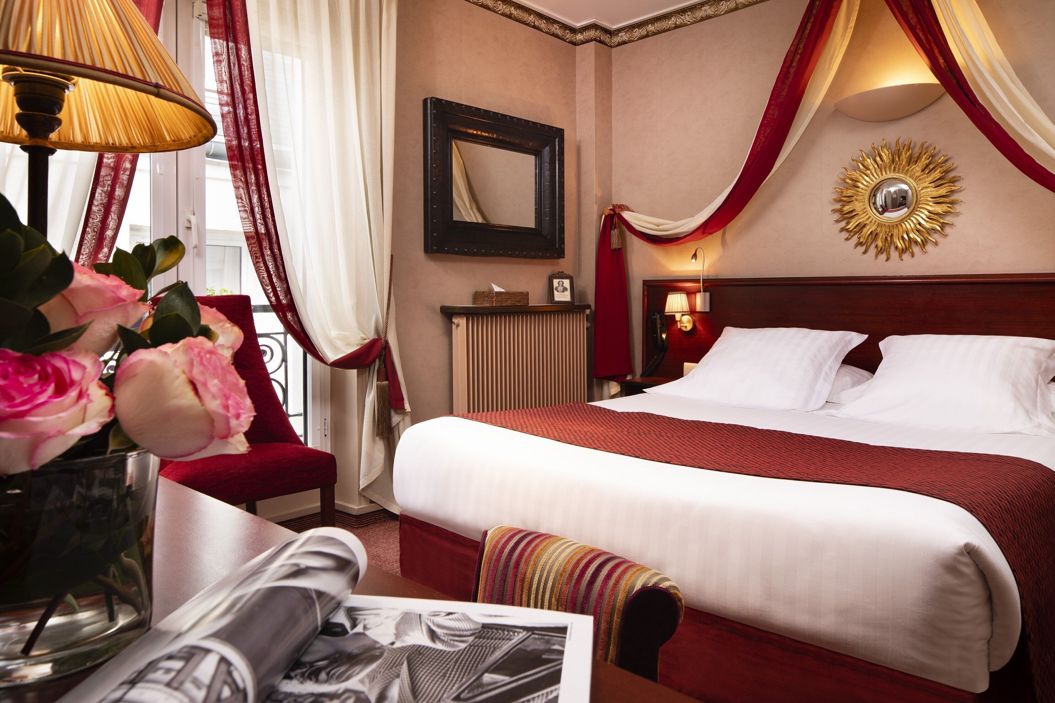 Habitación de hotel con una cama grande, una mesita de noche con lámpara, un jarrón con flores y una ventana con cortinas.