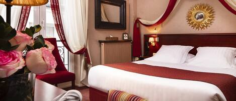 Habitación de hotel con una cama grande, una mesita de noche con lámpara, un jarrón con flores y una ventana con cortinas.