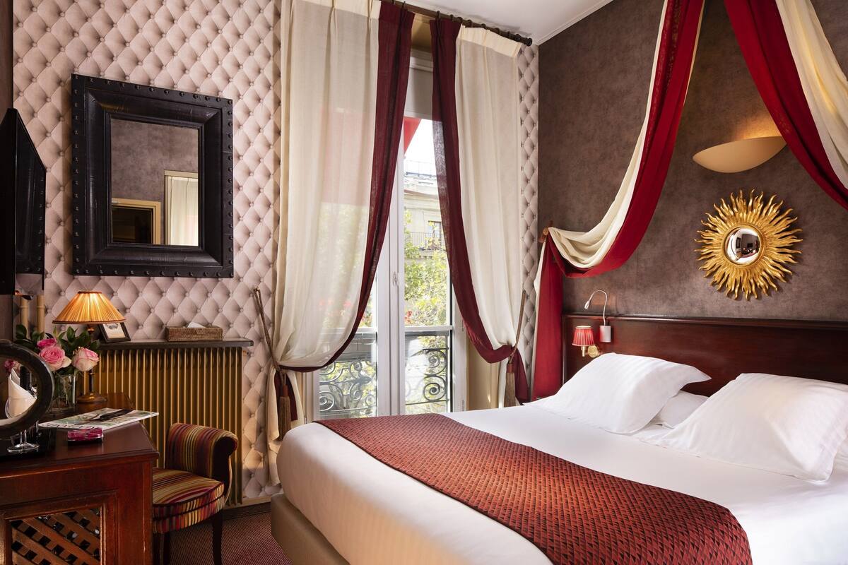 A hotel room with a large bed, a mirror, a lamp, a chair, and a window with curtains.