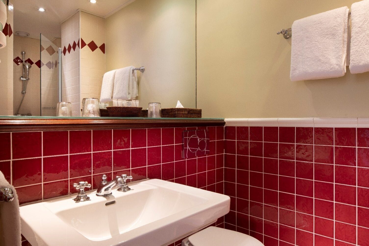 Un baño con lavamanos blanco, paredes con azulejos rojos, cabina de ducha de vidrio y un espejo sobre el lavamanos.