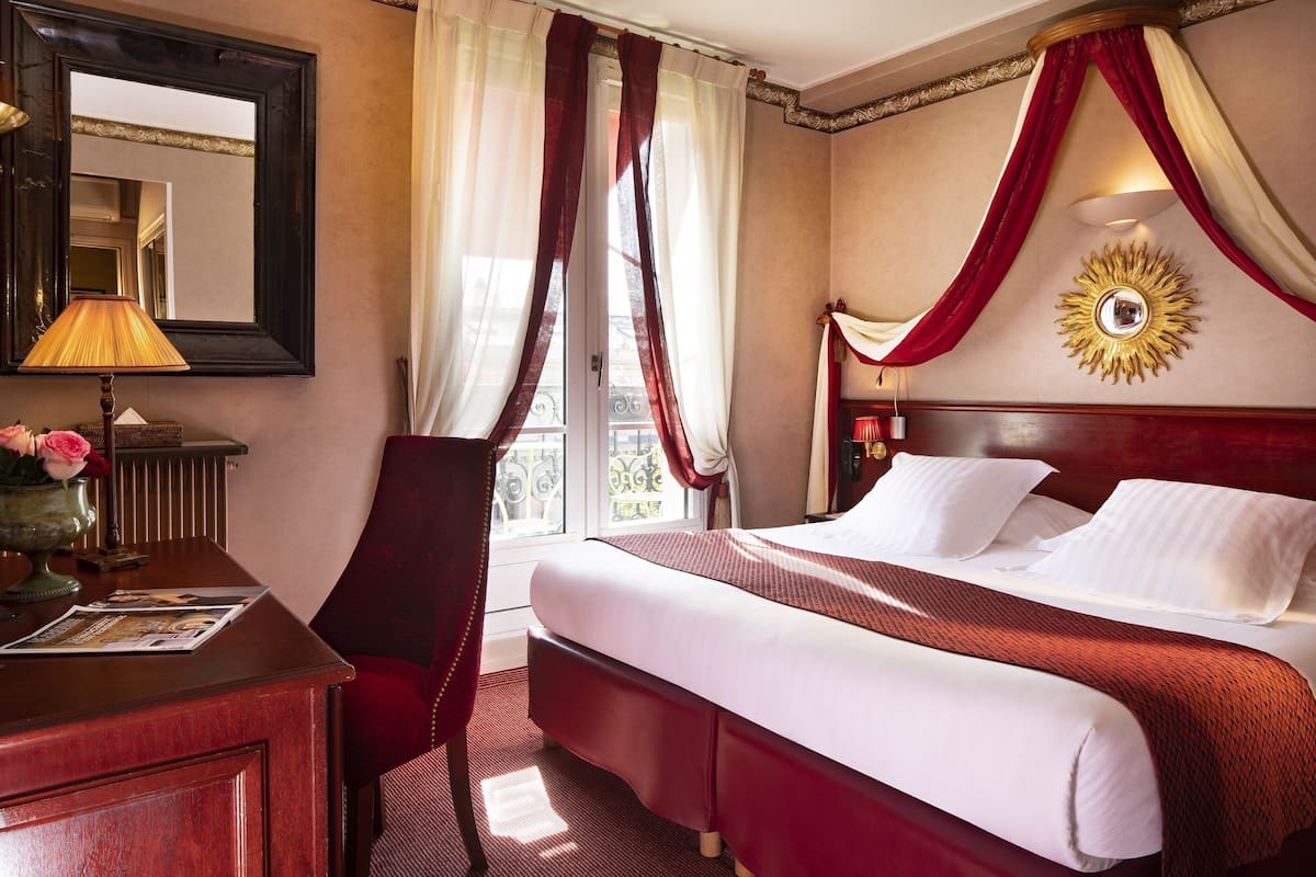 A hotel room with a large bed, a desk with a lamp, a chair, and a window with curtains.