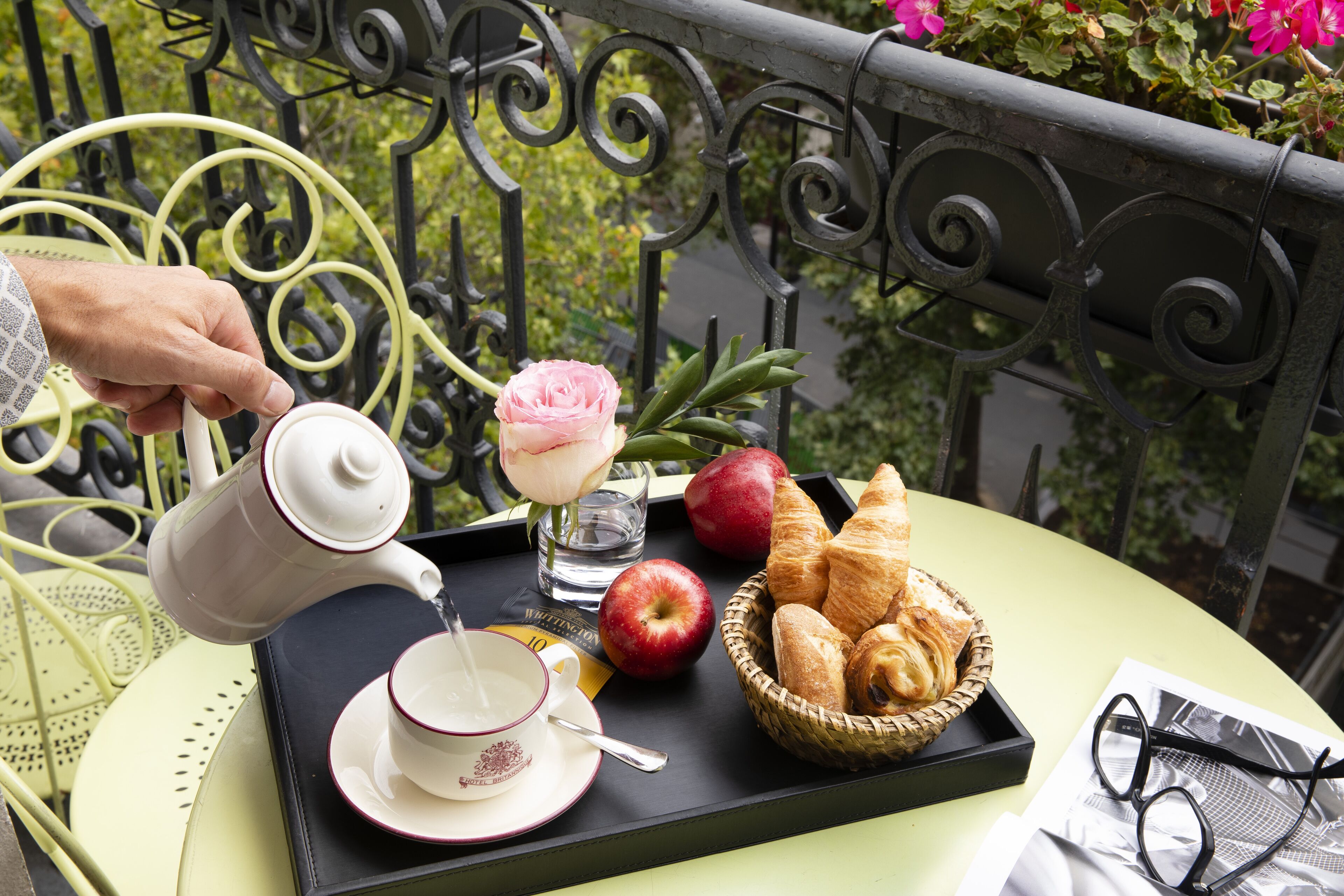 Een dienblad met een theepot, kopje, roos, appels, croissants en glazen op een balkon.