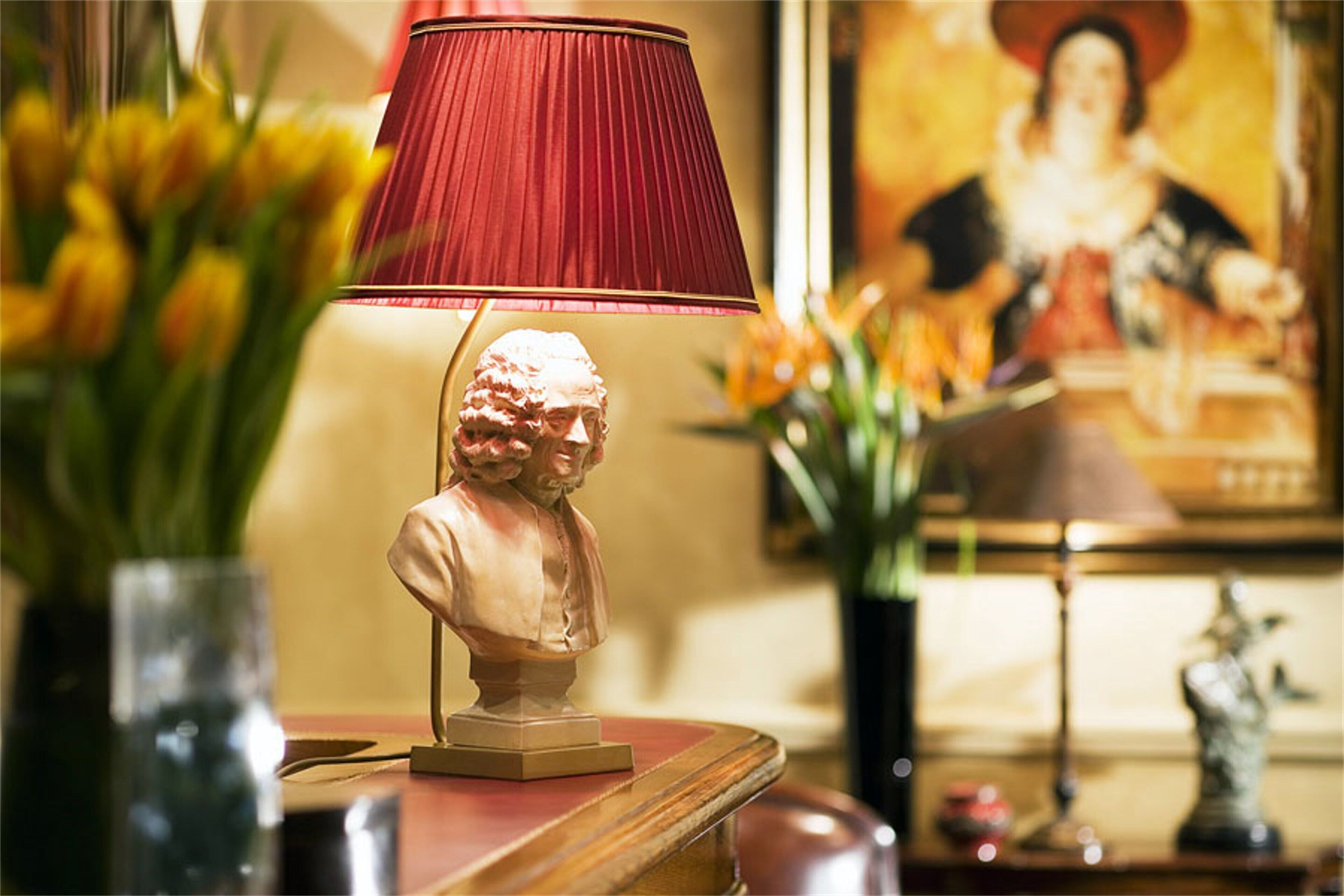 A table with a red lamp, a bust sculpture, and a framed painting in the background.